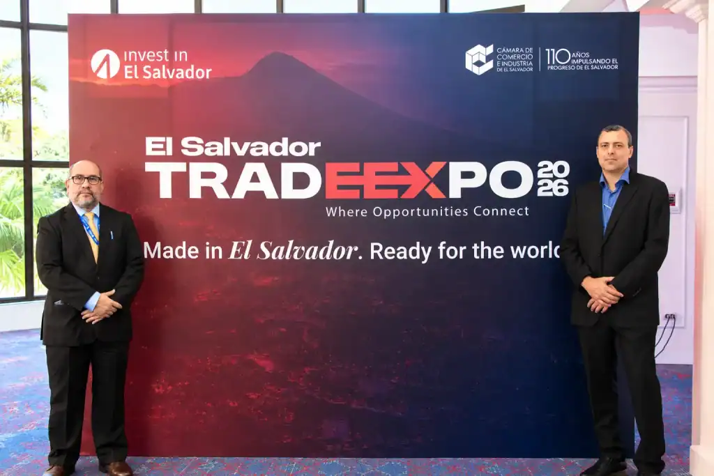 Dos hombres en traje formal posan a los costados de un gran panel promocional del evento 'El Salvador TRADEXPO 2026', el cual muestra el lema 'Made in El Salvador. Ready for the world' y logos de Invest in El Salvador y la Cámara de Comercio e Industria.
