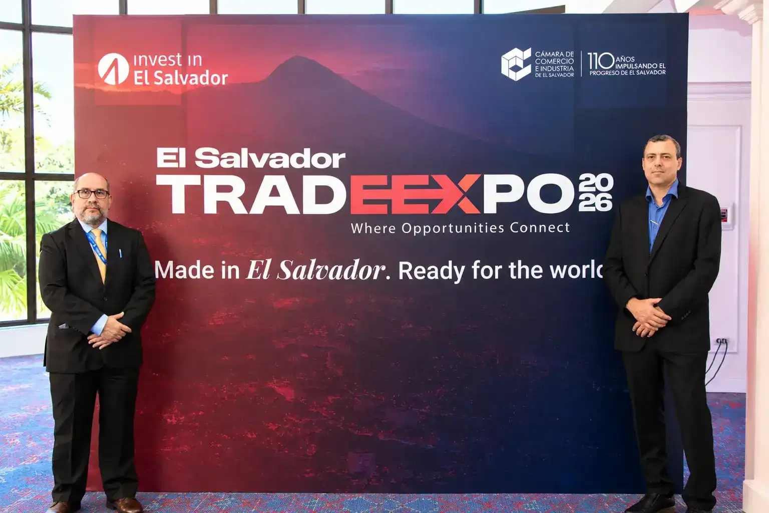 Dos hombres en traje formal posan a los costados de un gran panel promocional del evento 'El Salvador TRADEXPO 2026', el cual muestra el lema 'Made in El Salvador. Ready for the world' y logos de Invest in El Salvador y la Cámara de Comercio e Industria.